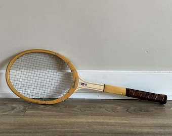 Könnte beinhalten: Ein Vintage-Tennisschläger aus Holz mit einem braunen Ledergriff. Der Schläger hat einen gelben und weißen Rahmen mit einem bespannten Schlägerkopf.