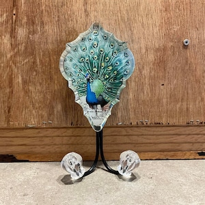 Op de afbeelding: Een decoratieve wandhaak met een pauwenmotief. De haak is gemaakt van metaal en heeft een transparante glazen voorkant met een vintage ansichtkaartbeeld van een pauw met zijn uitgespreide staartveren. De haak heeft twee punten om items aan op te hangen.