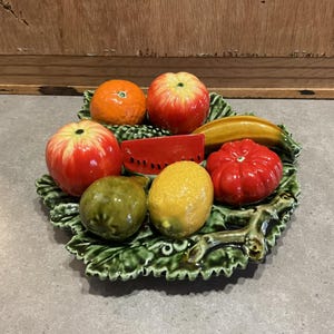May include: A ceramic fruit bowl with a green leaf design. The bowl features a variety of fruits including a red apple, a yellow lemon, a green pear, a red tomato, a yellow banana, a red watermelon slice, and an orange.