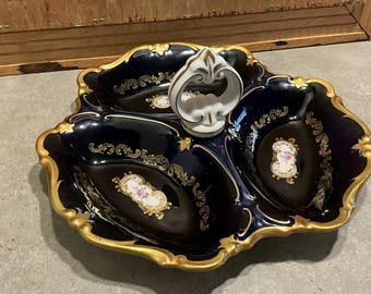 May include: A navy blue porcelain serving dish with three compartments and a white handle. The dish has a gold trim and floral accents. The handle is white with gold detailing. The dish is on a gray surface.