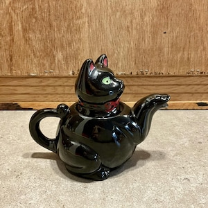 Op de afbeelding: Zwarte keramische theepot in de vorm van een kat met groene ogen en een rode strik. De theepot heeft een tuit en een handvat.