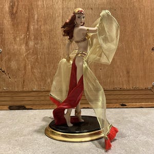 Puede incluir: Figura decorativa de una mujer con un traje dorado y rojo, con una diadema y joyas doradas. La figura está sobre una base negra con un borde dorado. La mujer posa con los brazos extendidos, sosteniendo la tela dorada.