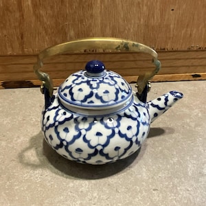 Puede incluir: Una pequeña tetera de cerámica con un diseño floral azul y blanco. Cuenta con un asa de latón y un pomo azul en la tapa. La tetera está diseñada con un pico para verter y probablemente se utiliza para servir té.