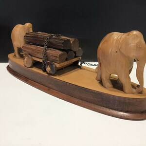 May include: A wooden sculpture of two elephants pulling a cart loaded with logs. The elephants are carved in a realistic style and the cart has wheels. The sculpture is mounted on a wooden base.