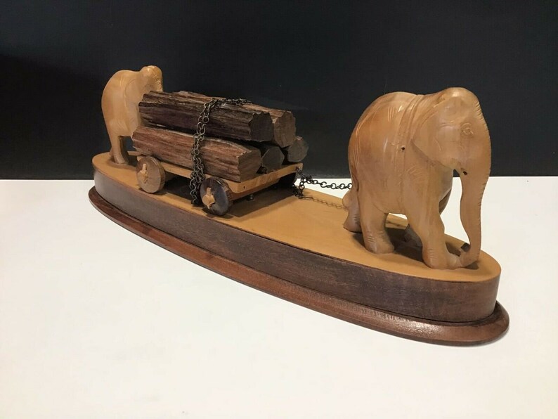 May include: A wooden sculpture of two elephants pulling a cart loaded with logs. The elephants are carved in a realistic style and the cart has wheels. The sculpture is mounted on a wooden base.