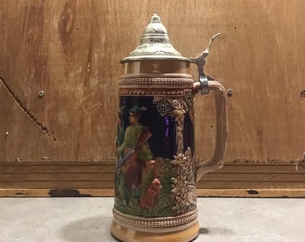 May include: A ceramic beer stein with a pewter lid and handle. The stein features a hunting scene with a man, a dog, and a forest background. The text "Gute Jagd" is visible on the stein.