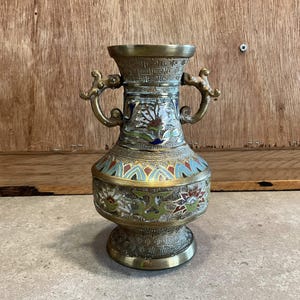 Peut inclure: Un petit vase en laiton avec deux anses et des motifs floraux complexes dans des tons de bleu, vert, rouge et blanc. Le vase présente un motif géométrique autour du haut et du bas.