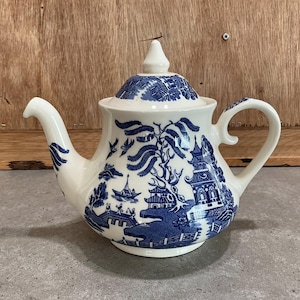 Könnte beinhalten: Eine weiße Keramik-Teekanne mit blauem Willow-Muster. Die Teekanne hat einen gebogenen Ausguss, einen abgerundeten Körper und einen passenden Deckel mit Knauf. Der Griff ist eine gebogene Schlaufe. Das Design umfasst Pagoden, Brücken und Bäume.