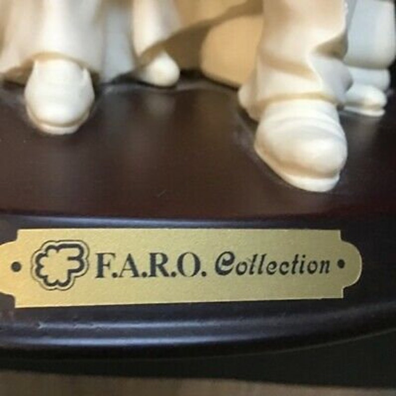May include: A white porcelain figurine of a group of people standing on a wooden base. The base has a gold plaque with the text "F.A.R.O. Collection."