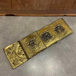May include: A set of four gold-toned metal wall plaques with embossed designs. Each plaque features a different scene with figures and intricate details. The plaques are connected together with hinges, allowing them to fold up.