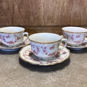 Könnte beinhalten: Drei weiße Teetassen und Untertassen mit rosa Rosenmustern und goldenen Verzierungen. Die Tassen und Untertassen sind auf einer grauen Oberfläche mit einem hölzernen Hintergrund angeordnet.