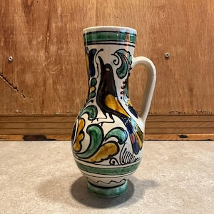 May include: A ceramic jug with a cream-coloured base featuring hand-painted designs of a black and yellow bird surrounded by green and yellow floral motifs. The vessel has a single handle, a flared rim, and horizontal green bands at the top and base.