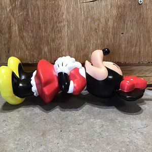 May include: A Minnie Mouse-shaped water bottle, featuring a red bow, black body, and yellow shoes. The bottle has a white straw and a red, polka-dotted lid. The figure is lying on a concrete surface with a wooden background.