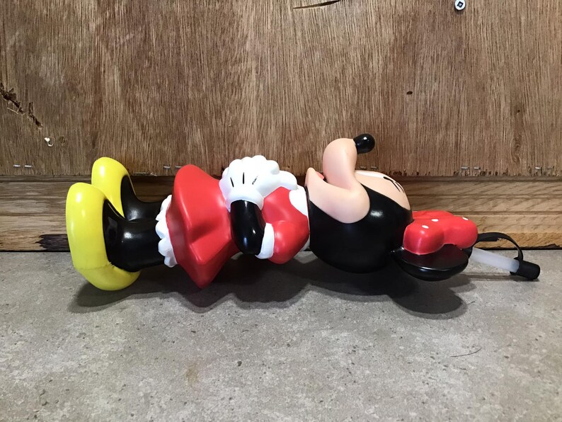 May include: A Minnie Mouse-shaped water bottle, featuring a red bow, black body, and yellow shoes. The bottle has a white straw and a red, polka-dotted lid. The figure is lying on a concrete surface with a wooden background.