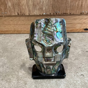 May include: A decorative mask with a geometric design, crafted from iridescent abalone shell pieces. The mask features eye and mouth openings and is mounted on a black square base. The shell's colors include blues, greens, and purples.