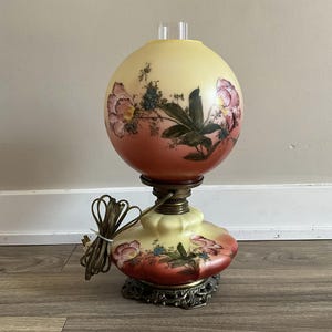 Peut inclure: Une lampe vintage en verre avec un motif floral peint à la main. La lampe a un globe rond, jaune et rouge avec un motif floral et une base dorée.