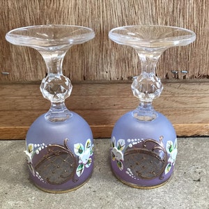 May include: Two clear glass stemmed goblets with a frosted lavender base. The goblets are decorated with gold and white floral designs.