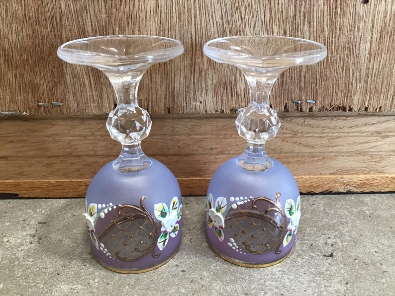 May include: Two clear glass stemmed goblets with a frosted lavender base. The goblets are decorated with gold and white floral designs.