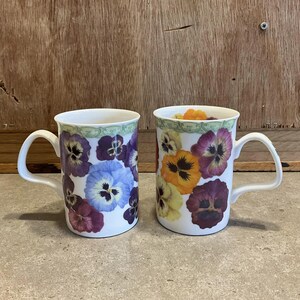 May include: Two white ceramic mugs with handles. One mug has a floral pattern of purple, blue, and yellow pansies. The other mug has a floral pattern of orange, yellow, and purple pansies.