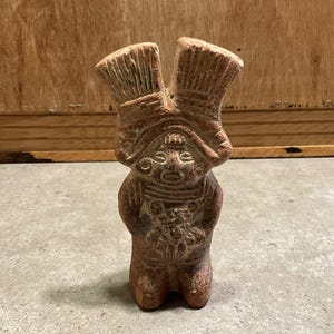 以下が含まれることがあります： 小さな赤褐色のセラミック製の置物。様式化された人物で、大きな頭飾りがあり、小さなオブジェクトを持っています。置物は、木製の背景のある灰色の表面に立っています。