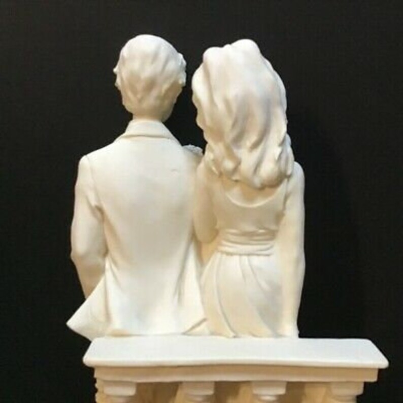 May include: White ceramic figurine of a couple, a man and a woman, standing side by side. They are both wearing simple clothing and have their arms around each other. The figurine is sitting on a white pedestal.