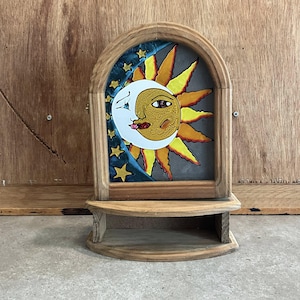 Puede incluir: Una estantería decorativa de madera con un diseño celestial. La obra de arte representa un sol y una luna sonrientes, con un fondo estrellado azul. La estantería es de madera clara y tiene una base curva.
