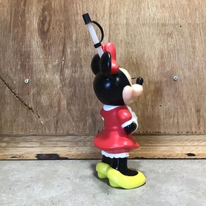 May include: A Minnie Mouse-shaped plastic tumbler with a black head, red bow, and red dress. The tumbler has a white straw and yellow shoes. The tumbler is standing on a gray surface against a wooden background.