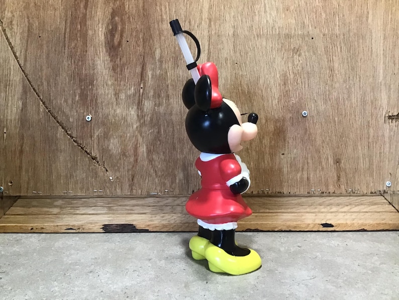 May include: A Minnie Mouse-shaped plastic tumbler with a black head, red bow, and red dress. The tumbler has a white straw and yellow shoes. The tumbler is standing on a gray surface against a wooden background.