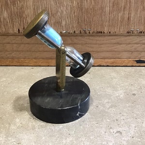 May include: A brass hourglass with blue sand on a black marble base. The hourglass is tilted to the side and the sand is flowing from the top to the bottom.