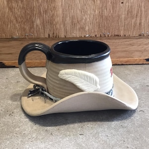 May include: A ceramic coffee mug shaped like a cowboy hat with a feather design. The mug is beige with a black rim and a red accent. The hat is beige with a black band.