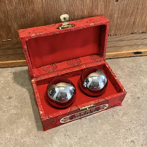 Peut inclure: Une boîte rouge ornée avec un fermoir doré, contenant deux boules métalliques brillantes de couleur argentée. La boîte est doublée de tissu rouge et présente un motif. Les boules reflètent l'environnement.