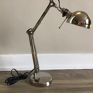 May include: A chrome desk lamp with an articulated arm and a circular base. The lamp has a domed lampshade and a black power cable. The lamp is designed for task lighting and is suitable for a desk or workspace.