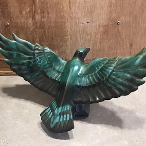 May include: A teal green ceramic sculpture of a bird with its wings spread wide. The bird is in flight and has a detailed design.