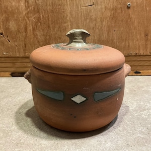 May include: A round, terracotta-coloured ceramic pot with a lid. The lid has a decorative pattern and a small handle. The pot features a design of blue and white shapes. Two small handles are on the sides.