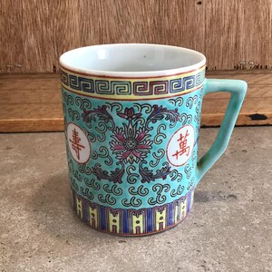 Può includere: Una tazza in ceramica turchese con un motivo floreale e caratteri cinesi. La tazza ha un bordo giallo e blu intorno alla parte superiore e inferiore.