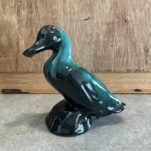 Könnte beinhalten: Keramik-Entenfigur mit glänzendem, dunkelgrünem und schwarzem Finish. Die Ente sitzt auf einem kleinen, dunklen Sockel. Die Figur hat einen langen Schnabel und steht.