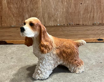 Peut inclure: Une figurine de chien cocker spaniel brun et blanc avec des oreilles tombantes. Le chien est debout sur ses quatre pattes et a une expression espiègle sur son visage.