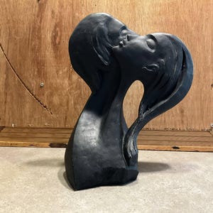 Op de afbeelding: Een zwarte sculptuur die twee figuren in een omhelzing afbeeldt, met hun gezichten dicht bij elkaar. De sculptuur heeft een gladde, matte afwerking en een vloeiend, abstract ontwerp. Het haar van de figuren is gedetailleerd en de sculptuur staat tegen een houten achtergrond.