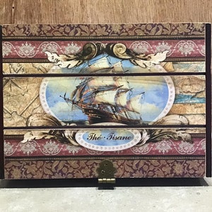 May include: A wooden box with a decorative design featuring a ship sailing on the ocean. The box has three drawers and a brass latch. The text "The Tisane" is printed on the front of the box.