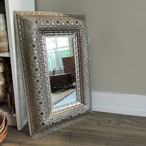 以下が含まれることがあります： 装飾的で質感のあるフレームが付いた、銀色の長方形の鏡。 フレームはモザイクのような模様と、縁に沿った装飾的な透かし彫りが特徴です。 鏡は、茶色の革のバッグとラグのある部屋を映しています。