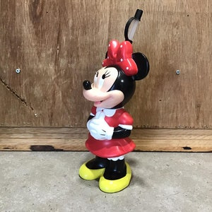May include: A Minnie Mouse figurine, featuring a black head and ears, a red dress with white accents, and yellow shoes. The figurine has a red bow with white polka dots and is standing on a gray surface.