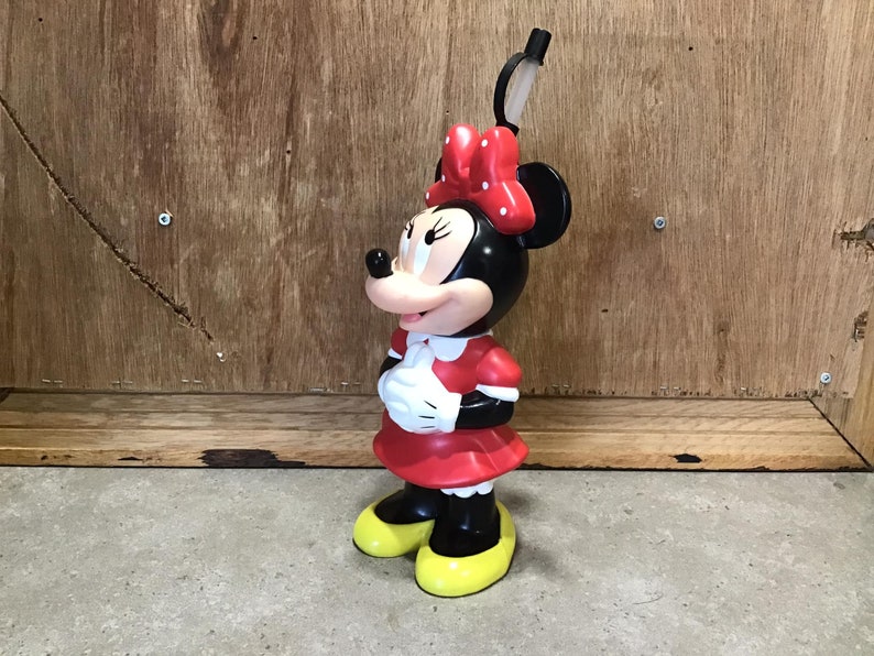 May include: A Minnie Mouse figurine, featuring a black head and ears, a red dress with white accents, and yellow shoes. The figurine has a red bow with white polka dots and is standing on a gray surface.