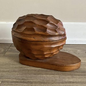 Puede incluir: Una caja de madera tallada a mano con tapa, con forma de nuez. La caja es de color marrón cálido y se asienta sobre una base de madera a juego. La tapa tiene un patrón geométrico texturizado.