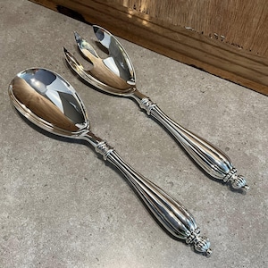Könnte beinhalten: Ein Paar silberne Salatbestecke. Eines ist ein Löffel, das andere eine Gabel mit drei Zinken. Beide haben geriffelte Griffe mit dekorativen Details und ein kronenartiges Design am Ende. Das Besteck liegt auf einer grauen Oberfläche.