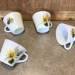 Pode incluir: Quatro ch&aacute;venas de ch&aacute; de cer&acirc;mica branca com desenhos florais amarelos e brancos. As ch&aacute;venas t&ecirc;m uma forma arredondada e uma pega no lado.