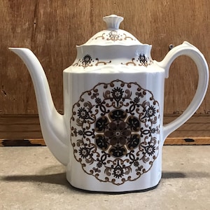 May include: White ceramic teapot with a brown and gold floral design. The teapot has a lid and a handle.