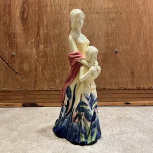 Può includere: Una statuetta in porcellana color crema che raffigura due figure, una in piedi dietro l'altra con le braccia che la avvolgono. Le figure indossano lunghe gonne fluenti decorate con dettagliati motivi di fiori di iris blu e verdi e uno scialle rosa.