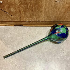 May include: A colorful glass plant watering globe with a long, slender stem. The globe is a mix of green, blue, and red glass pieces. The stem is a dark green color. The watering globe is designed to slowly release water into the soil.