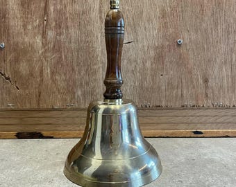 May include: A brass bell with a wooden handle. The bell is polished and shiny. The handle is dark brown with a light brown grain.