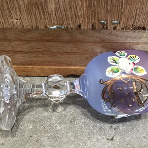 May include: A purple glass goblet with a gold rim and hand-painted floral design. The goblet has a clear glass stem and base.